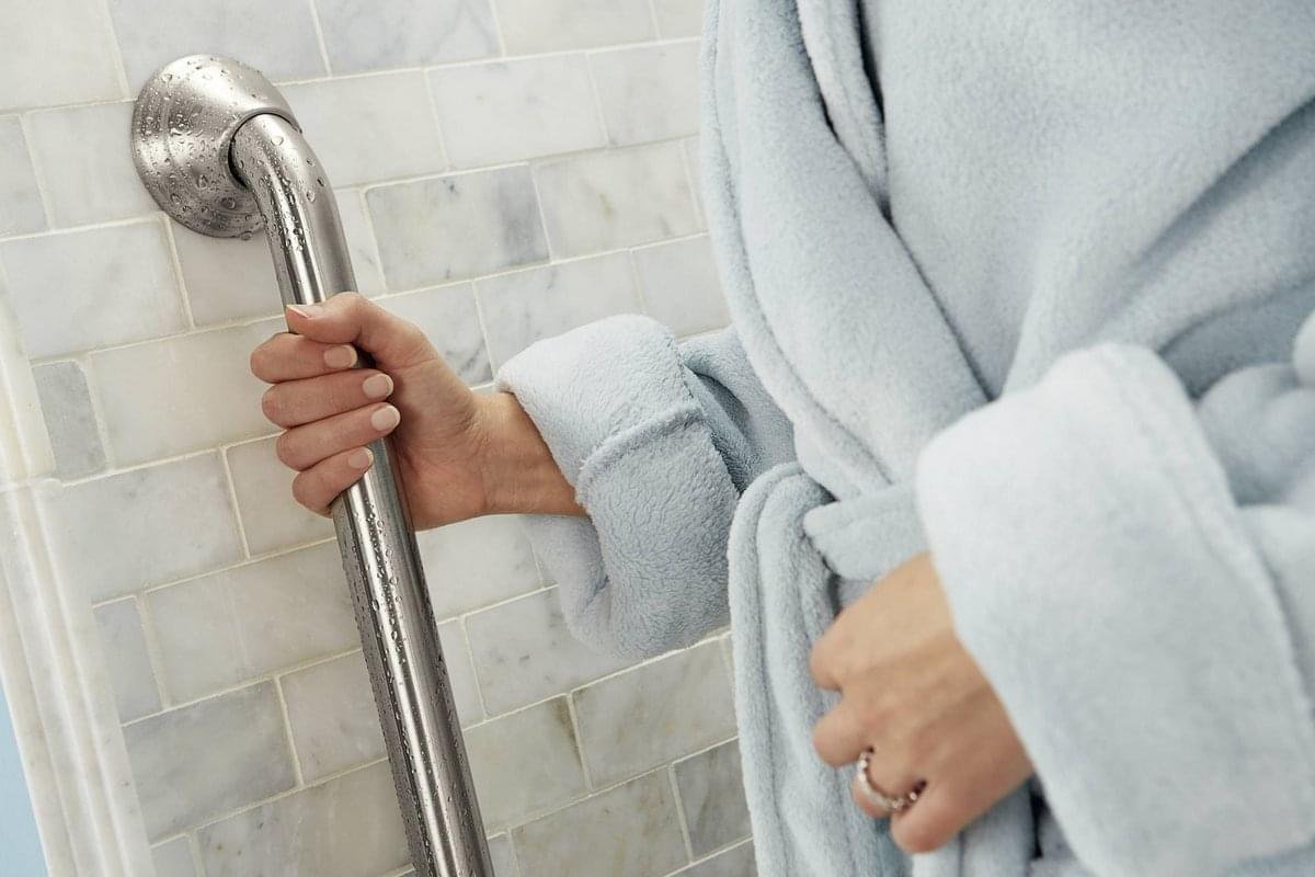 Person in a light blue bathrobe holding a stainless steel grab bar in a bathroom with marble tile walls
