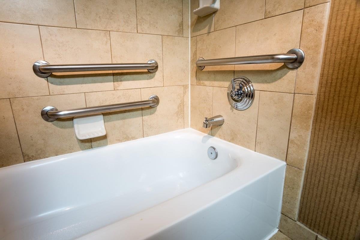Bathroom with a white bathtub and three stainless steel grab bars installed on beige tiled walls for safety and support.